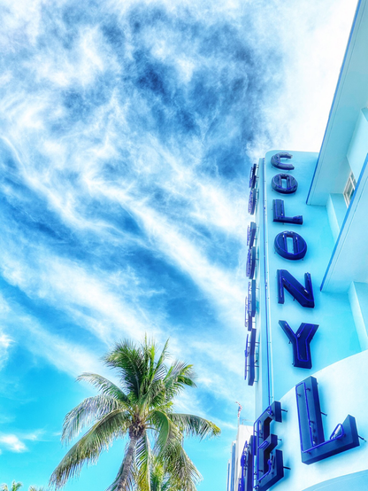 The blue sign of Hotel Colony on light blue building. Background is blue sky with clouds