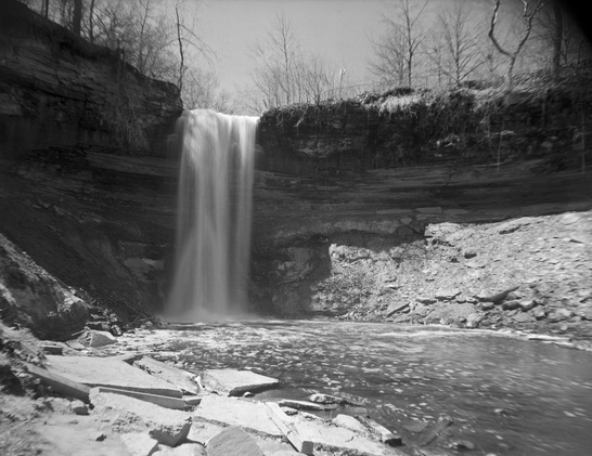 Narrow waterfalls photographed from within a deep gorge into a wide creek below. The scene is captured with a long exposure that produces a smooth effect on the water’s flow.