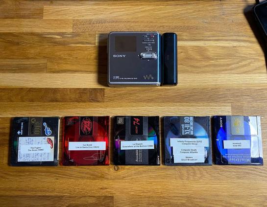 A silver, rectangular Sony Minidisc recorder on a wooden desk. Below it, five Minidiscs in various colours are pictured:
The Fugees -The Score (1996)
Car Bomb - Live in Santa Cruz (2023)
La D