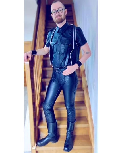 A young man wearing black leather boots, jeans, shirt, vest and bracers standing on a flight of stairs showing off his new leather jeans.