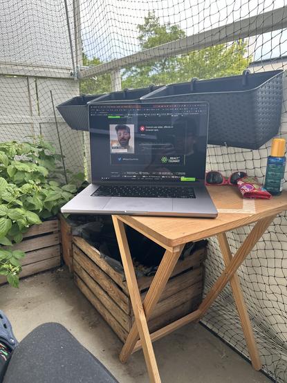 Picture of my laptop on a small table, outside at my balcony. On the screen is the react summit livestream, with the summit track. On the table there is also a bottle of sunblocker, Gummi bears and sunglasses. 

In the foreground you see my leg with a black trousers. In the background you see green plants and flower pots.