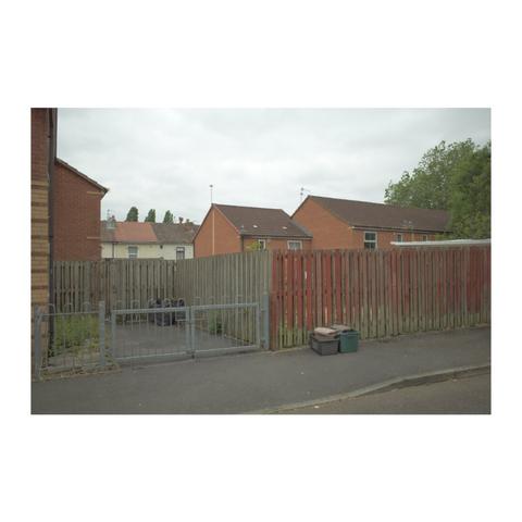 A narrow Street behind some red brick houses, with a rundown wooden fence and rubbish bins.