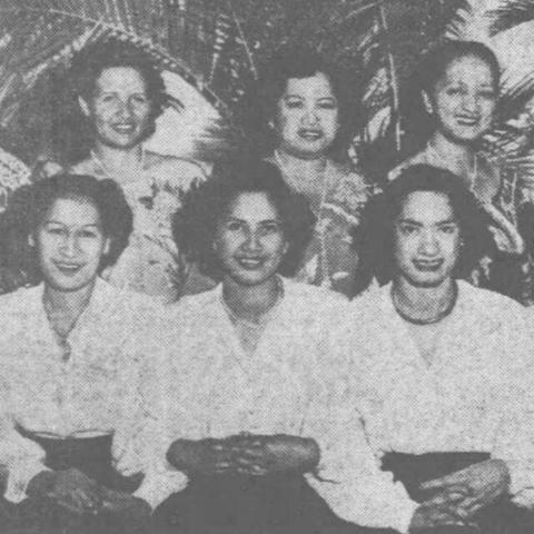 6 Hawaiian ladies doing music are smiling into the camera, 3 in front row in white blouses, 3 in back row in flowery ones.