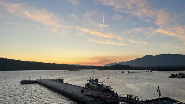 Looking to the West from a height. In the foreground is a large pier jutting diagonally to the left from the bottom right. There is a tugboat moored to it on the far side near the ramp to shore. Beyond is the harbour, glittering silver in the fading light of the day. There are some dark outlines of piers and booms and posts scattered in the water. Beyond the water is another thin dark outlin of a shore. And the horizon with orange highlighted smoke clouds low in the middle hugging the mountain range to the left and fasting to to yellow and blue sky above with some clouds.