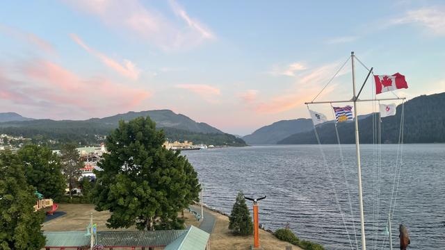Looking south down the Alberni Inlet from a height. In the foreground is a large tree shading a grassy area beside a gravel path that runs along the shoreline. There are some other some trees and bushes nearer the shore. Beyond is the harbour withheld disturbed wavy water in. The wind. There is a three lined sailing style flagpole to the left with three flags. In the far distance are three mountain ridge lines layered one after another on each side ending at the narrow channel of water.
