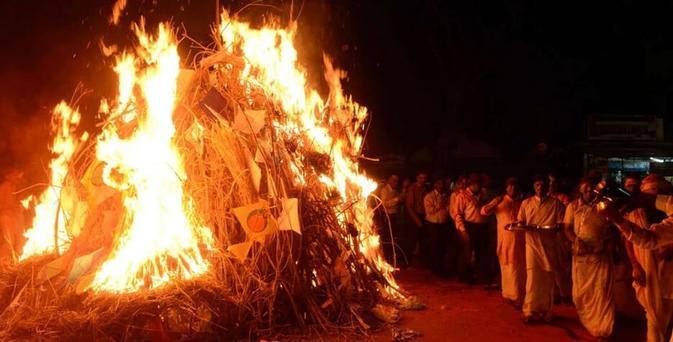 A large bonfire stacked with thin branches, twice as tall as the people gathered around it, burns ferociously as two lines of men walk around it in a large circle. Those closest to the camera are wearing white robes and carrying puja thalis. 

Holika Dahan symbolizes the burning of Holika, sister of asuran King Hiranyakashipu, during Satya Yuga. Hiranyakashipu thought of himself as a god and wanted everyone to worship him alone, putting to death any who refused. But after being influenced by the teachings of a sage while in the womb, his own son, Prahlada, became a devotee of Lord Vishnu. Outraged, Hiranyakashipu tried to kill Prahlada many times but each time Prahlada was protected by Vishnu's power. On one occasion Hiranyakashipu plotted to kill Prahlada by trapping him on a pyre, while under the protection of a boon his sister Holika had been granted by Brahma, and then exposing him to the flames. But the boon had been granted on the condition that Holika not use it to cause harm, and this condition combined with Vishnu's grace Prahlada was saved while Holika instead burned in the fire. Hiranyakashipu then tried to kill Prahlada himself once more, but Vishnu manifested as an incarnation of Narasimha and killed Hiranyakashipu instead. 

For this reason Holika Dahan celebrates the triumph of good over evil, and observing it is believed to banish fear and worry as well as offering relief from unfavourable planetary influences.