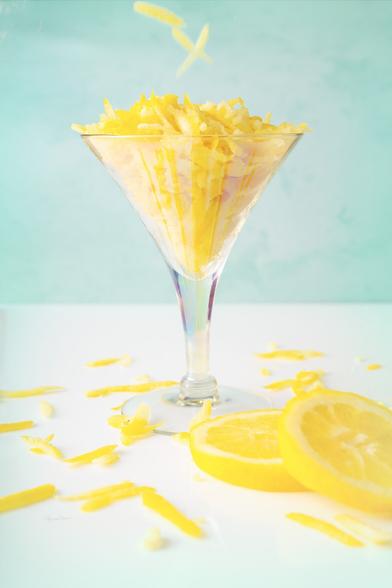 A fancy cocktail glass with a stem is full of lemon zest. Lemon zest is falling from above and lies all around. There are two lemon slices in the foreground.