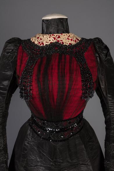 The bodice of a black and red gown trimmed with white lace at the very top. The bodice is draped in sheer tulle and covered with black beads. The shoulders have a slight puff. The satin is moiré so it has a water-like pattern to it.