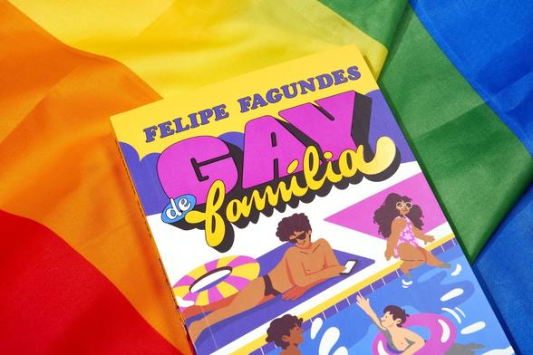 Foto colorida do livro Gay de Família, de Felipe Fagundes, disposto sobre uma bandeira nas cores do arco-íris, a bandeira LGBTQIAPN+. Na capa do livro há uma ilustração de um rapaz e uma garota com trajes de banho, à beira duma piscina.