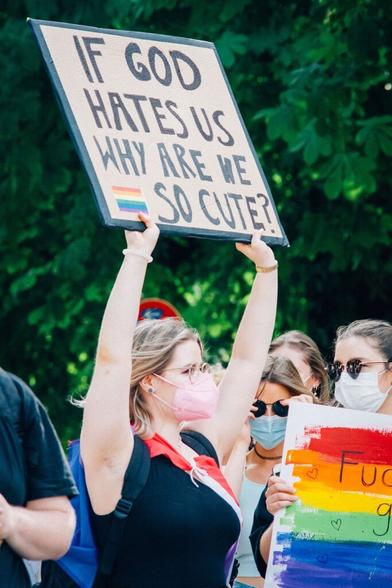 A person wearing a flag wrapped round their shoulders holding a sign in a crowd that reads "If god hates us why are we so cute?" with a rainbow flag on the sign.