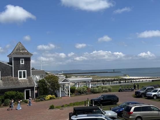 The photo shows the Atlantic Ocean on the horizon. In front of it is a beach and a gray Cape cod cottage house. The sky is blue with puffy clouds. In front of the cottage a group of brides maids walk