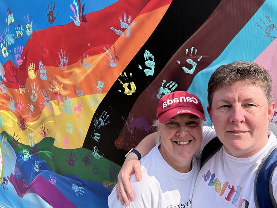 Selfie of Jen and Sarah in front of the Pride banner aka the handprint wall.
