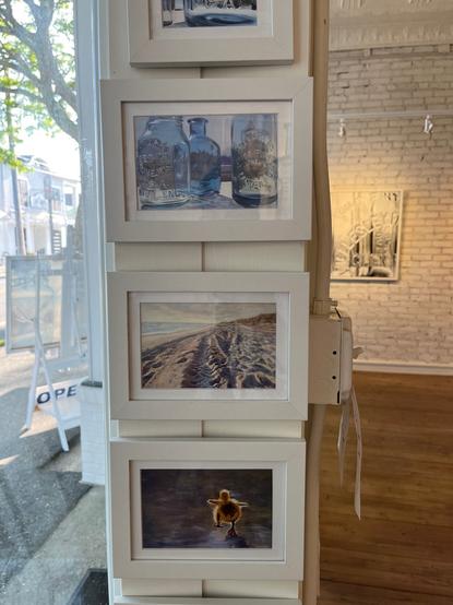 The photo shows a wall in an art gallery. Hanging on the wall are paintings of water glasses, the ocean and a baby duck