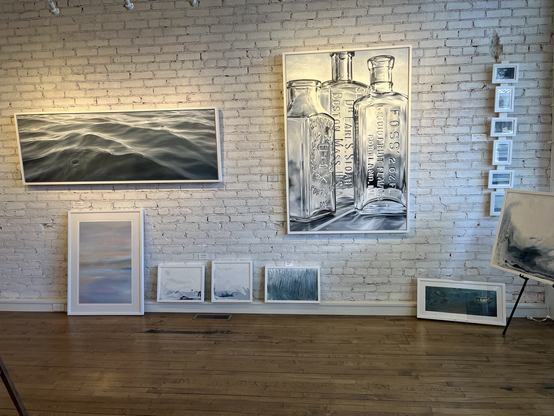 The photo shows the wall of an art gallery. The wall is white. Hanging on the wall are paintings of glass bottles and the ocean