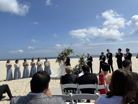 The photo shows a bride and groom and their wedding party getting married on a breach. In the background is the Atlantic Ocean. The bridesmaids are to the left and are wearing blue dresses and holding flowers. To the right are the groomsmen who are wearing black suits and clapping as the bride and groom embrace