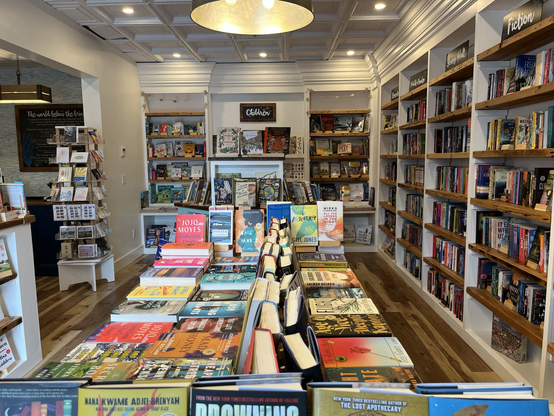 Photo shows a bookstore. Immediately ahead is a table top covered in books. There are wall to wall bookshelves full of books