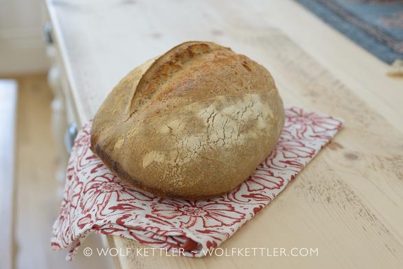 The photograph shows a rustic Mediterranean inspired bread displayed on the wooden top of a traditional farmhouse table.