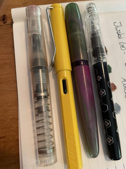 4 fountain pens, a clear pen with visible mechanism, a bright yellow pen, a purple and green marble pen and a black pen with a geometric flower design.