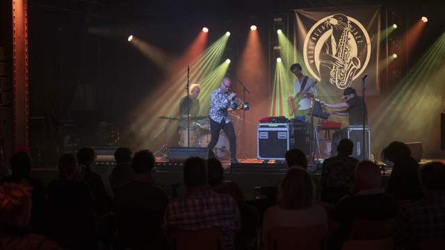 Band onstage, with drums, trumpet, bass guitar and keyboards, with red and green stagelighting and a festival logo backdrop, in front of an audience in the dark.