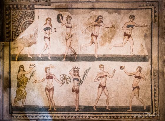 A top down view of a gymnasium floor mosaic depicting ten women performing different activities or sports; some are running, some are playing catch, some are lifting weights.