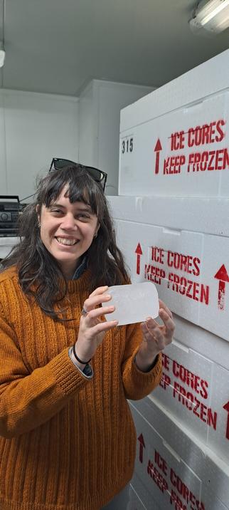 A woman smiling into camera holding a cylinder of ice! She's standing next to boxes that say "ice cores keep frozen"