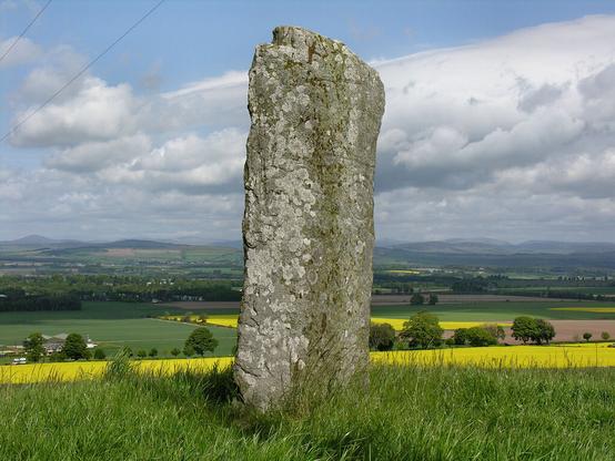 The Keillor Pictish symbol stone. The image shows a grey carved stone in the centre of the frame. It is covered in lichen, but traces of carvings can be made out in it. The stone is set on a hillside that descends behind it past agricultural field to distant hills. The scene is in sunlight.