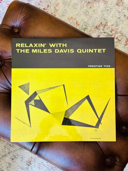 Photo of a record album jacket laying on a tan leather stool. 

The jacket is cubist figure of a woman, all triangles cut in crisp black, against a vivid yellow backdrop. 

A black banner runs across the top of the jacket where, in yellow, text reads, ‘Relaxin’ with the Miles Davis Quintet’.