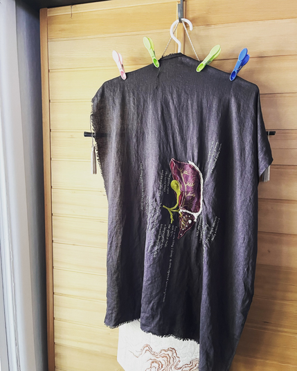 Lia’s completed liver embroidery hangs on a white plastic hanger on the side of a wooden sauna. The light from the window is bright but orangey.