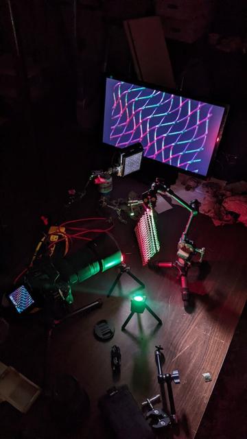 Photo of a lighting setup on a tabletop. The camera is aimed at a wavy metal grilling mesh. It is suspended by a posing arm at a diagonal angle, and lit with 3 LED lights of varying colors and sizes. The camera output is shown on a monitor in the background.