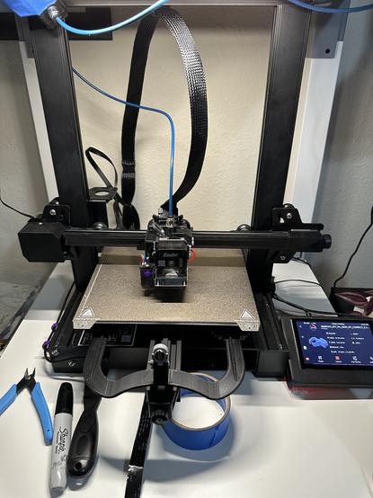 A 3D Printer sitting on a small table illuminated my bright LED lighting. In Front of the printer are various tools like flush cutters, scraper, and blue tape. Also in the picture is the screen for the printer displaying the current print status and a small representation of the model being printed.