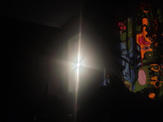 Morning light creeping through the crack of an open window and the window frame, creating a star-shaped light pattern, mostly vertical, scattering to the sides. The room is dark, due to underexposure, only showing a glimpse of the head of a guitar, tuning keys lighting up slightly, on the left. On the right, curtains lit up by the sun light from the outside, showing a psychedelic pattern of organic shapes in blue, pink, red, orange and green.