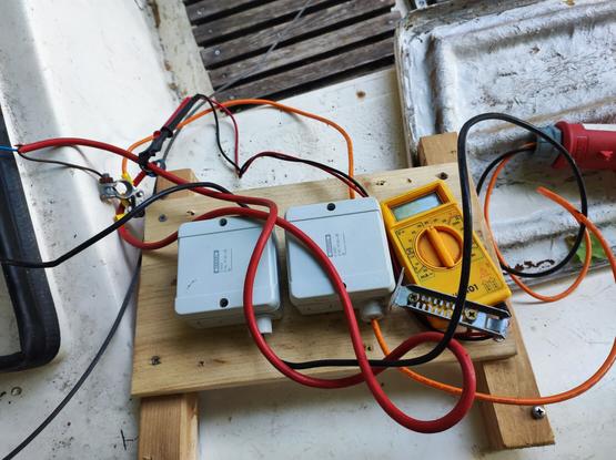 Ein Blick auf die "Elektrik". Aus zwei Feuchtraum Verteilerdosen kommen diverse Kabel. Ein gelbes Multimeter ist mit einem Stück Blech und zwei Schrauben aus ein Holzbrett geschraubt. Die Prüfspitzen sind mit Klebeband an weiteren Kabeln befestigt.