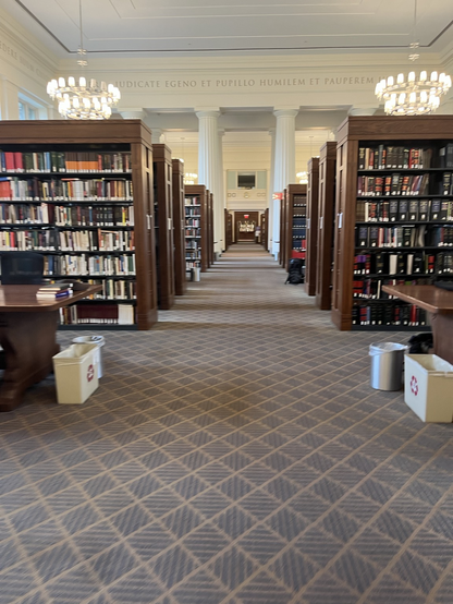 The photo shows a great hall with 2 stacks of books on either side of a center walk way. The great hall is Harvard law library
