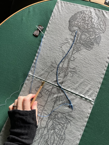 An image of an old engraving of a vagus nerve is printed on sulky material and basted to green cotton in a huge wooden embroidery hoop. Some stitching of one of the nerve lines has been done in blue chain stitch in perlé cotton. Lia's hand can be seen next to the stitching holding a tambour hook.