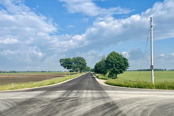 The junction of a gravel road (out of frame, a foreground element not visible) arriving from low-left-and-right at a road of freshly-paved blacktop spread beneath us and arrowing ahead toward the far horizon between rows of trees in summer green—it narrows and fades in the same direction we are cycling. The mottled cumulus typical of a cool near-summer day hangs overhead giving the sky a pleasing texture. In the foreground, the pavement is patterned with tire tracks of dust laid down by vehicles passing through. Fields lie either side of the journey ahead, the fresh spring crops a lighter green than the trees.