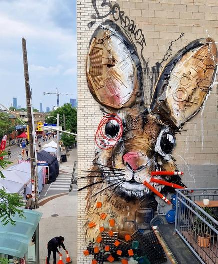 Installation. An art installation on the beige, partially tiled exterior wall of a multi-story building. The image of a happy looking beige rabbit has been assembled from garbage and sprayed around. Carton pieces, red wires, orange plastic containers and pilons and many other materials have been combined with painted pieces to create a wonderful mural.