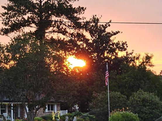 A typical American home, view if the front yard below an orange sky with the glowing sunrise peeking from behind distant treetops