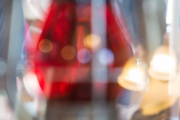 A photograph of a railroad lantern with red glass in a museum exhibit. The photograph is deliberately blurred. Reflected circles of light cross the display glass in the center of the frame - orange, yellow and blue, and the reflection of an antique wall light with fluted glass shades is visible to the right. The whole image is lit by bright sunlight coming through the windows.