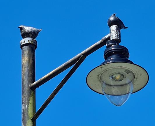 Two aluminium pigeons perched on top of a street light against a clear blue sky.