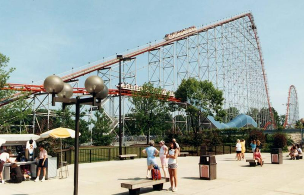 A view of a train on Magnum XL-200 ascending the first lift hill.  There is also another train on the brake run, returning to the state.  In the foreground, guests wait and watch as the train gains altitude.  The first and second hills can be seen in the image, as well as the final tunnel and brake run.