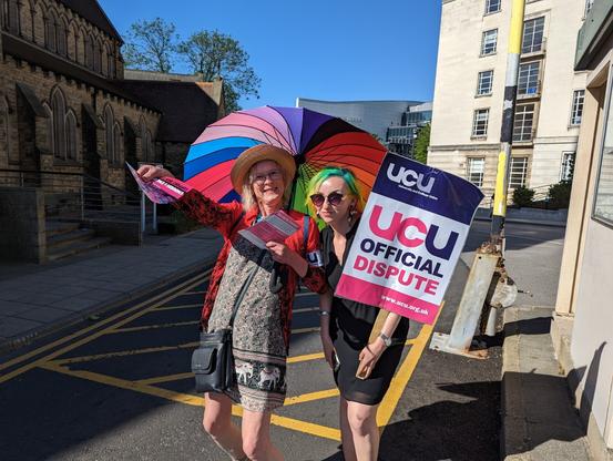 Two smiling pickets, one with rainbow hair the other with a rainbow umbrella, in the sunshine at University of Leeds main entrance