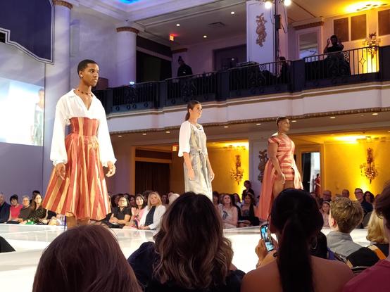 Three models on a raised runway showing off my sister's "Lumen" collection. Featuring a masculine model wearing a dress with vertical red and gold stripes and a white v-necked top with puffy sleeves, a feminine model wearing a vertical-striped blue and gold jumpsuit with white sleeves underneath, and a plus-sized feminine model wearing a dress with the same red and gold striped fabric but arranged with different sections at different angles forming a chevron pattern. Other fashion students and faculty can be seen looking at the models from beyond the runway