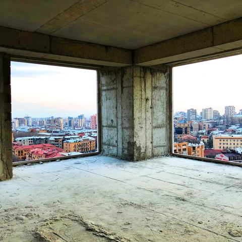 A corner of a high-rise building under construction is shown. The ceiling and columns are all exposed concrete. Two window wells frame either side of a beam in the center of the picture and in the distance you can see many other building towers and rooftops in hues of orange, red, gray, and beige.