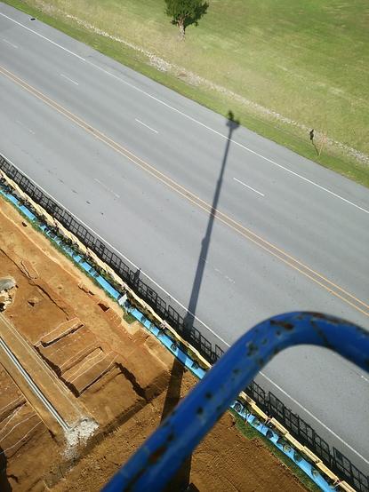 Photograph showing a lawn (top right), a road (center, running from the top left to the bottom right corner) and the excavated survey area (lower left), with the shadow of a 32 m high aerial work platform with me inside.