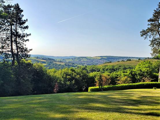 Standing on a mown lawn with an evening's long shadows from trees across it looking out across Cotswold hills with a blue summer sky above.