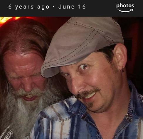 Two men facing the camera. The one in front has a mustache and cap and is looking scared. The man in back is grey bearded with eyes closed about to yell.