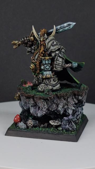 Dwarf paladin miniature standing on grassy outcrop with a small cave underneath wearing stone armour, carrying a sword and hammer with tiny insects and animals around