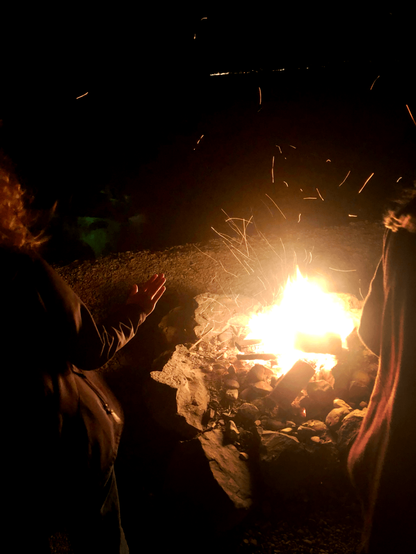 A woman warming herself up by the fireplace at night. The scene is happening at the side of the lake with the lights of the cities on the other side of the lake visible far in the distance.