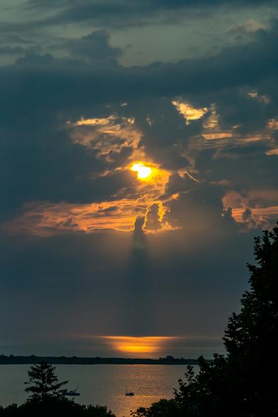 A color photograph, from a high vantage point, looking out across a harbor and towards Cape Cod Bay. The sun is well above the horizon, but because of an overlapping set of clouds, it still looks like dawn. The sun glows were visible, illuminating the clouds surrounding it. A darker band of clouds seepages that for the water and horizon. An orange reflection from the sun is visible on the water aimed towards the viewer. The outline of trees, shadowed and lower on the hill, frame the photograph at the bottom. A single boat sits at anchor in the harbor.