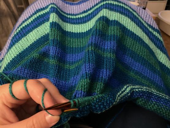HĂ€nde halten Stricknadeln, mit denen diese eine Decke aus Blockstreifen in blau und grĂŒn Tönen stricken.
Die Streifen sind unterschiedlich breit und folgen keinem erkennbarem Muster.
Die Decke liegt der strickenden Person auf den Beinen und bedeckt diese.